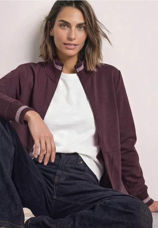 Frau mit kastanienfarbenem Blouson, weißem T-Shirt und Jeans sitzt lässig und blickt selbstbewusst.
