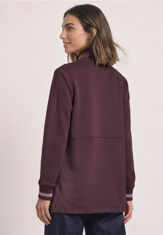 Eine Frau mit schulterlangem Haar trägt eine dunkelviolette Jacke und steht mit dem Rücken zur Kamera.