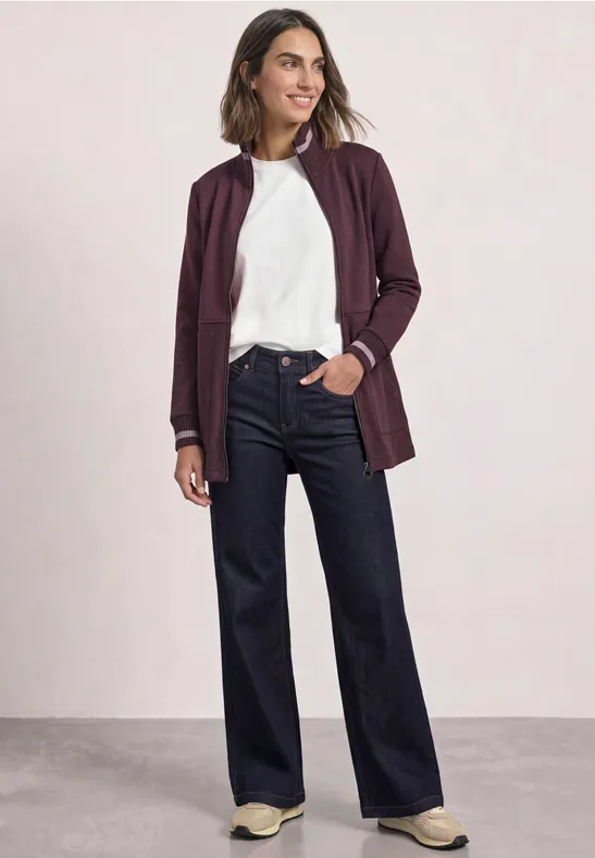Frau in weißem Shirt, bordeauxfarbener Jacke und weiten, dunklen Jeans, sportliche Sneaker.