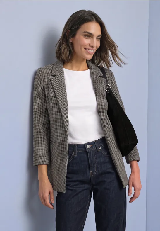 Eine lächelnde Frau trägt grauen Blazer, weißes Shirt, Jeans und schwarze Schultertasche vor hellem Hintergrund.