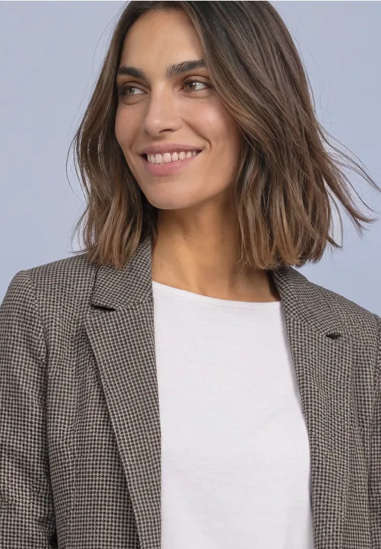 Eine lächelnde Frau mit mittellangen braunen Haaren trägt ein weißes Shirt und einen graubraunen Blazer.