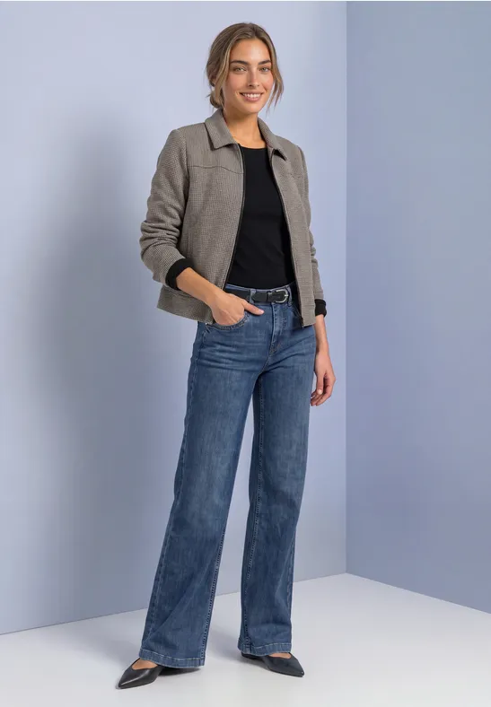 Frau in einer grauen, kurzen Jacke, schwarzem Top und weiten blauen Jeans vor hellem Hintergrund