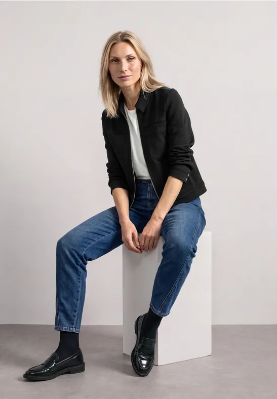 Eine blonde Frau sitzt auf einem weißen Würfel, trägt schwarzes Sakko, weißes Shirt, blaue Jeans und schwarze Stiefel.
