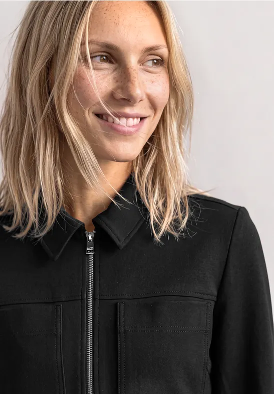 Nahe Aufnahme einer lächelnden Frau mit blonden Haaren in schwarzer Jacke, die seitlich in die Ferne blickt.