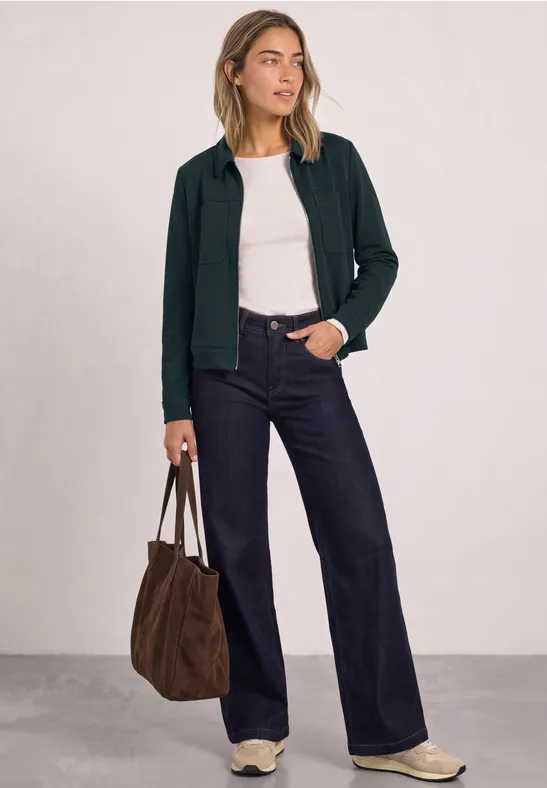 Frau in weißem Top, dunklem Cardigan, weiten Jeans, Sneakern, mit brauner Tasche vor neutralem Hintergrund.