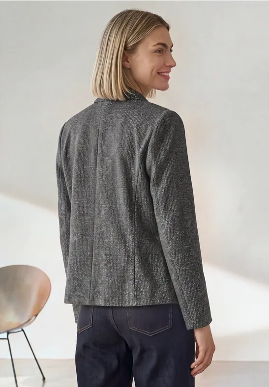 Frau mit blondem Bobhaarschnitt trägt grauen Blazer, steht im Profil, lächelt zur Seite.
