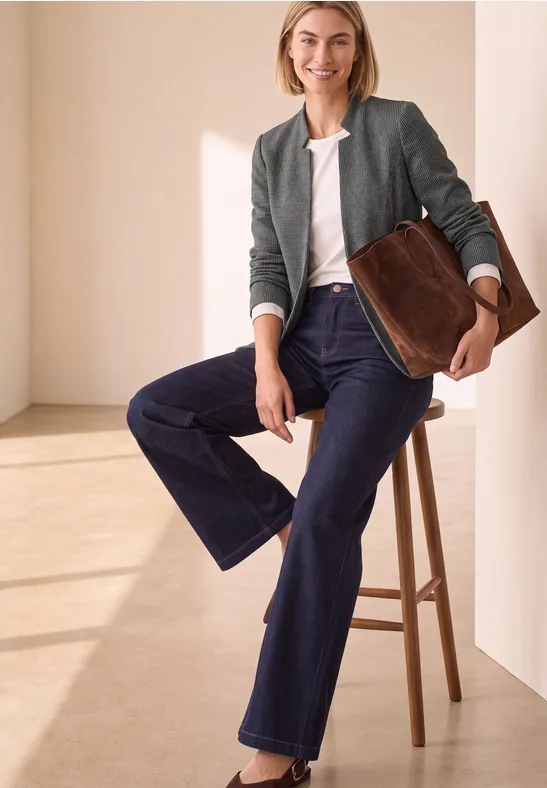 Eine Frau in weiten Jeans und Jacke sitzt auf einem Hocker, hält eine große braune Tasche.