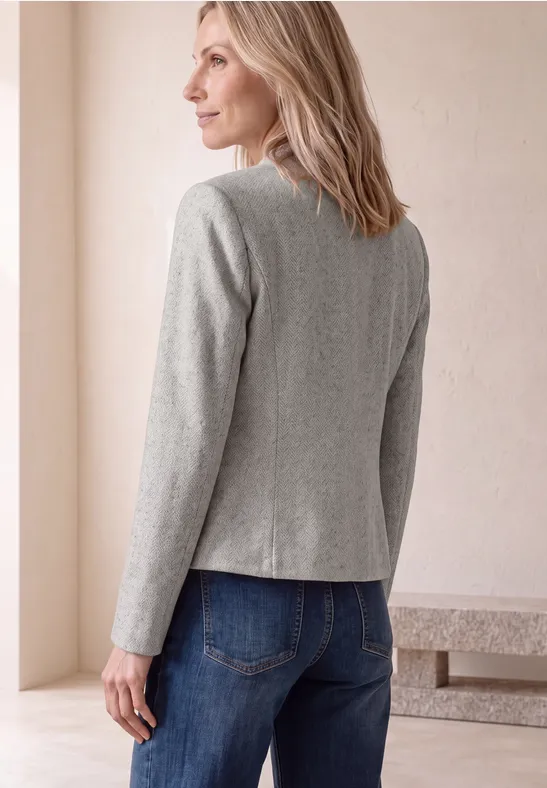 Frau von hinten in grauem Blazer und blauen Jeans vor hellem Hintergrund.