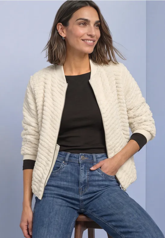Frau in weißer gesteppter Jacke, schwarzem Top und Jeans, lächelt vor hellem Hintergrund.