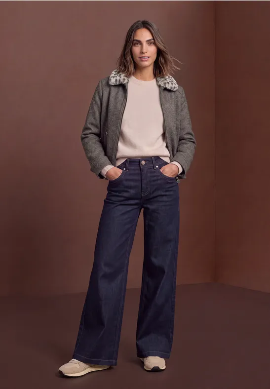 Frau in olivgrüner Jacke mit Pelzkragen, beigem Pullover, weiten Jeans und Sneakern vor braunem Hintergrund.