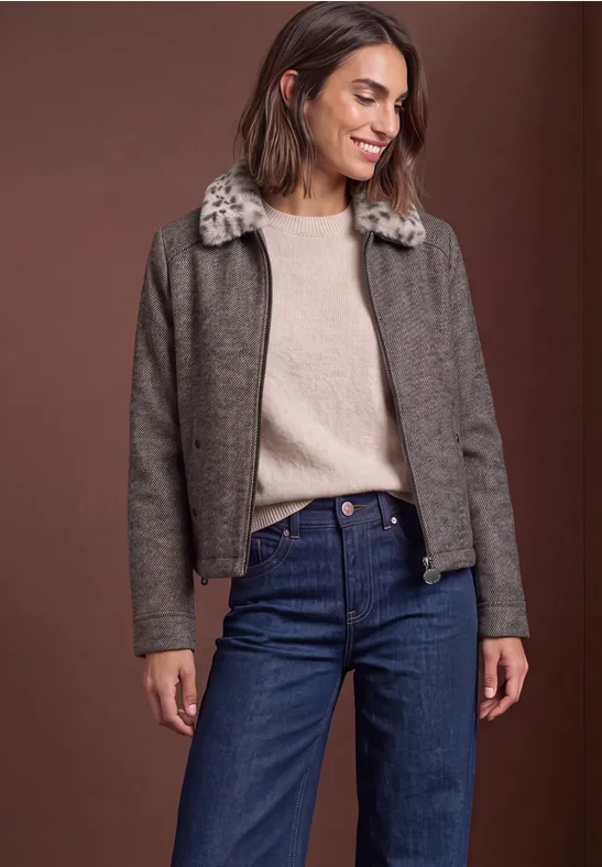 Frau in grauer Jacke mit Fellkragen, beigem Pullover und Jeans vor braunem Hintergrund.