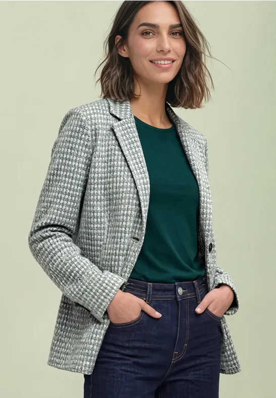 Femme portant une veste pied-de-poule, un t-shirt vert et un jean, les mains dans les poches, souriante.