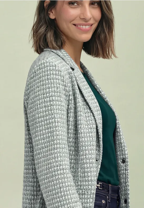 Une femme souriante en blazer pied-de-poule et haut vert foncé devant un fond clair