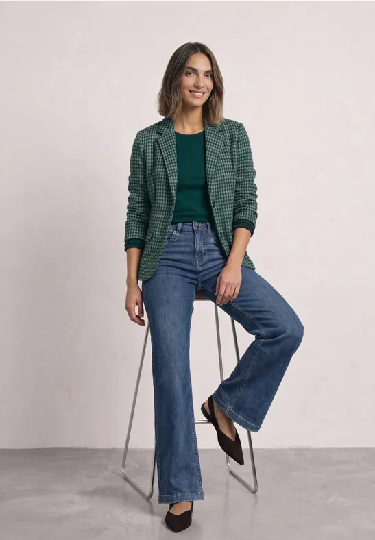 Femme assise sur tabouret, veste verte, haut vert, jeans larges, chaussures noires, souriante.