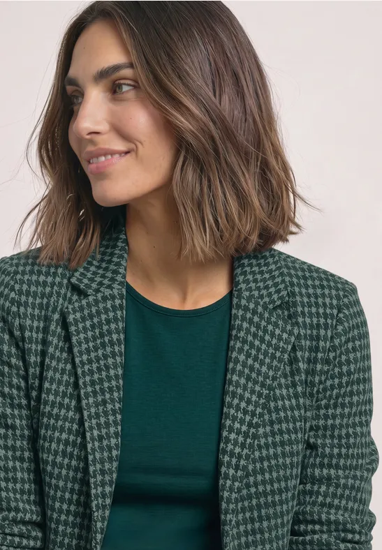 Femme brune aux cheveux mi-longs, souriante, portant un t-shirt vert et un blazer à motif pied-de-poule.