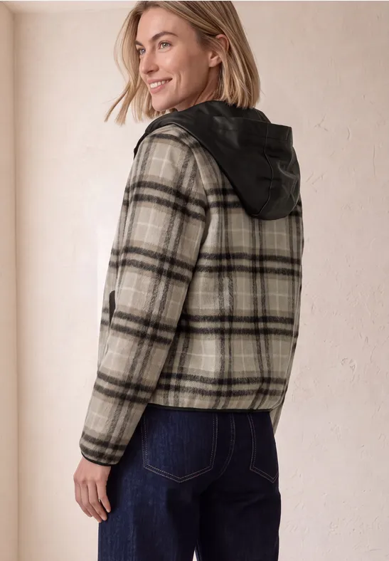 Frau in karierter Jacke mit Kapuze und Jeans, lächelnd über die Schulter blickend vor heller Wand.