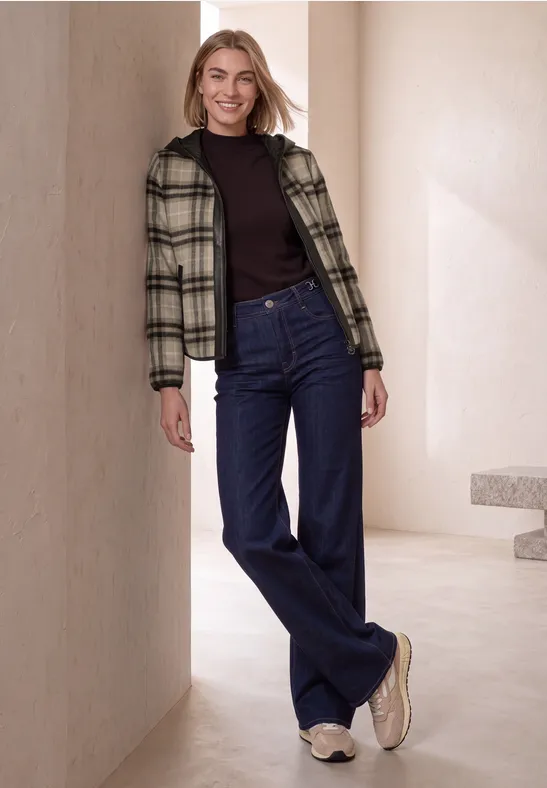 Modell in minimalistischer Umgebung lehnt lässig: karierte Jacke, schwarzer Pullover, weite blaue Jeans, weiße Sneaker.