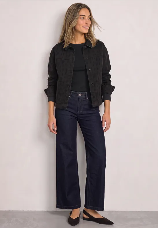 Frau mit blonden Haaren trägt schwarze Jacke, Shirt und weite Jeans vor hellem Hintergrund.