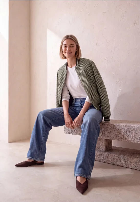 Frau sitzt lässig auf einer Steinbank, trägt Jeans, weißes Shirt und grünes Sakko, lächelt in minimalistischem Raum.