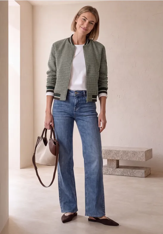 Frau in grüner Bomberjacke, weißem T-Shirt, Jeans und Loafers trägt Tasche in minimalistischem Ambiente.