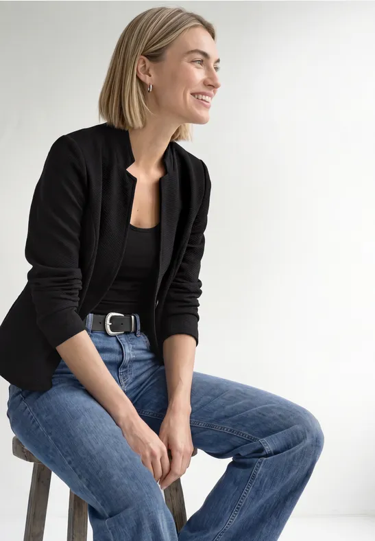Frau in schwarzem Blazer und Jeans sitzt lächelnd auf einem Hocker vor hellem Hintergrund.