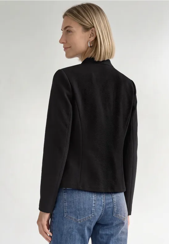 Frau mit kurzem blondem Haar in schwarzem Blazer und blauen Jeans, seitlich von hinten vor weißem Hintergrund.