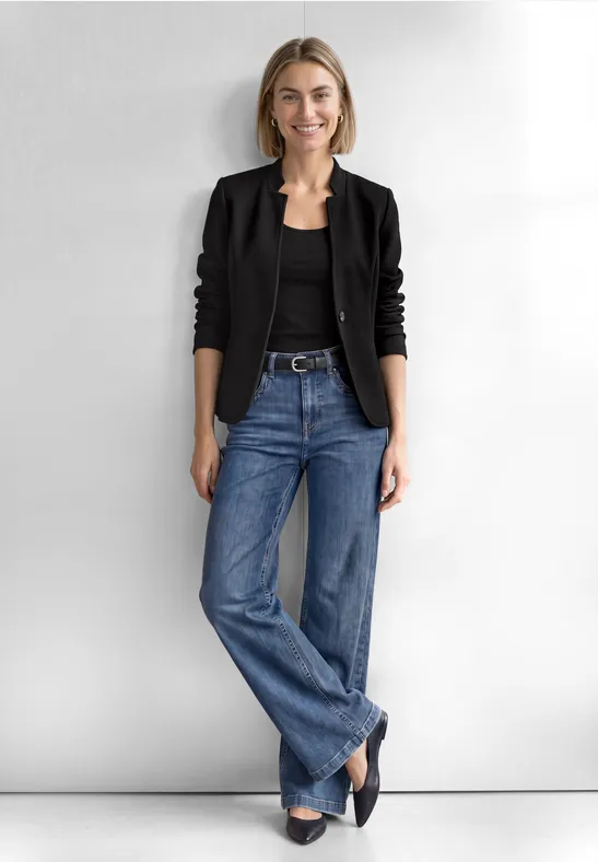Eine Frau steht vor hellem Hintergrund in schwarzer Jacke, schwarzem Top, weitem Jeans und schwarzen Schuhen.