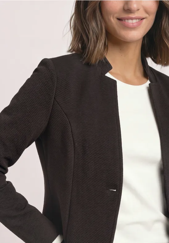 Frau trägt dunkelbraunen Blazer über weißem Top, lächelt leicht.