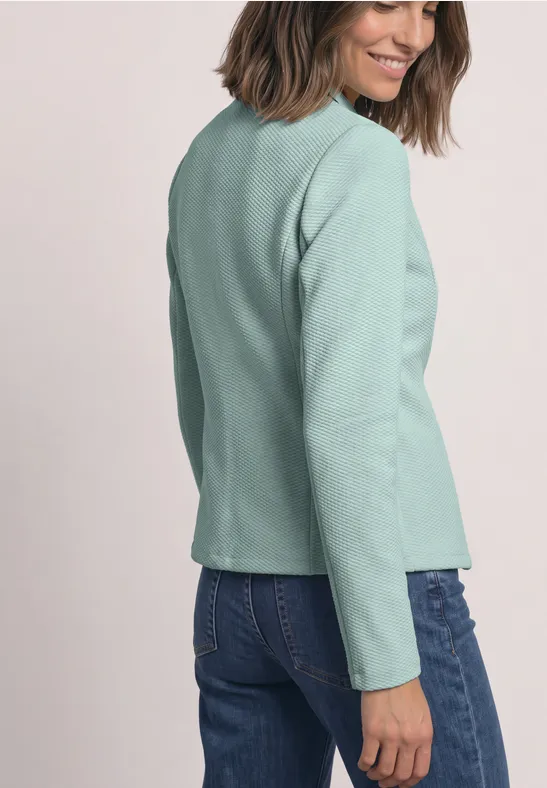 Frau in hellem grünen Blazer und Jeans, seitlich stehend und leicht lächelnd vor neutralem Hintergrund.