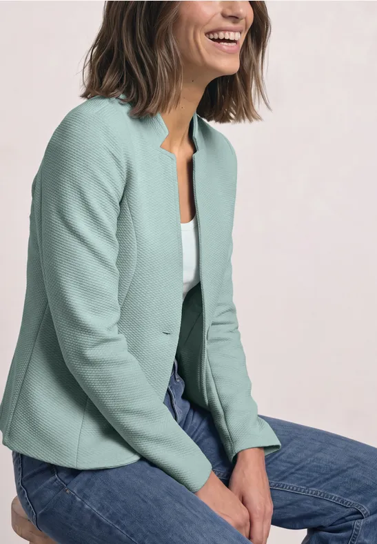 Eine lächelnde Frau sitzt mit verschränkten Händen auf dem Schoß in Jeans und mintgrünem Blazer.
