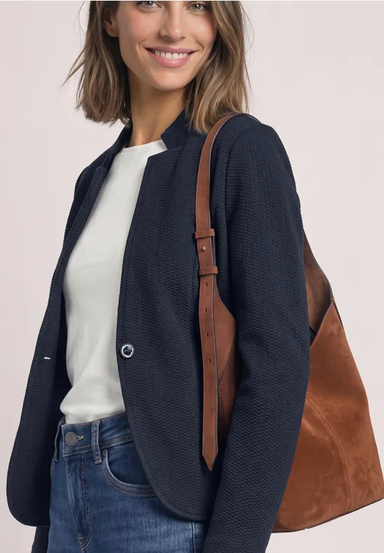 Eine lächelnde Frau in Jeans, weißem Shirt und dunkelblauem Blazer trägt eine braune Tasche.
