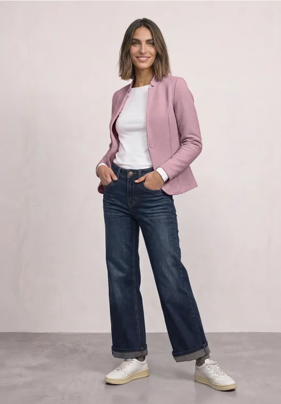 Frau in rosa Blazer, weißem T-Shirt, geraden Jeans und weißen Sneakern, lässig mit Händen in den Taschen stehend.