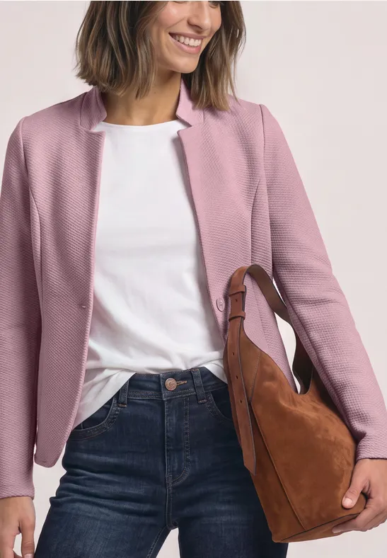 Frau trägt rosafarbene Jacke, weißes T-Shirt, blaue Jeans und hält eine braune Schultertasche.