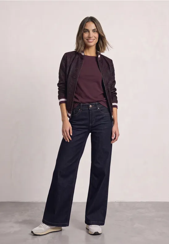 Frau in bordeauxroter Bluse, violetter Bomberjacke, weiten Jeans und weißen Sneakern.