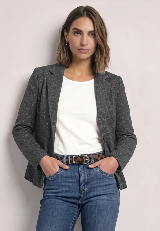 Frau in Jeans, weißem T-Shirt und grauem Blazer steht lässig mit Händen in den Taschen.