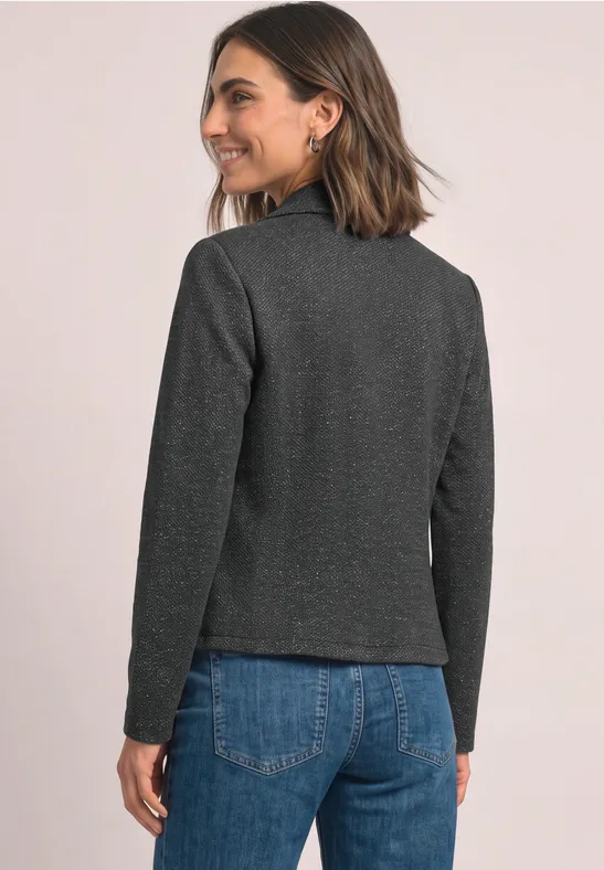 Frau in grauer Jacke und blauer Jeans, blickt über die Schulter, lächelnd.