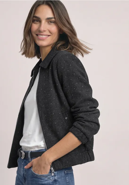 Eine lächelnde Frau mit braunen Haaren trägt eine schwarze Strickjacke und weißes Shirt.