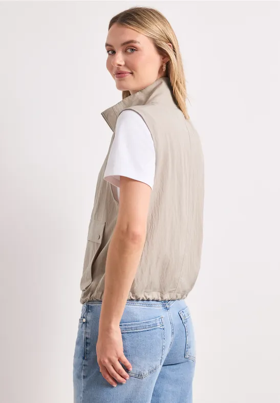 Zij-aanzicht van persoon in lichtbeige mouwloos jack, wit T-shirt en spijkerbroek, witte achtergrond.