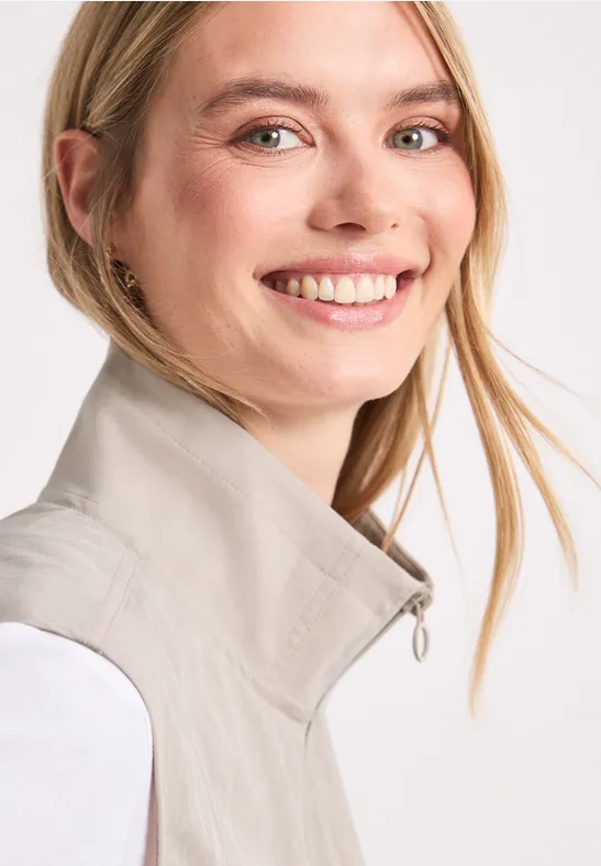 Een glimlachende vrouw met blond haar draagt een licht gilet voor een witte achtergrond.
