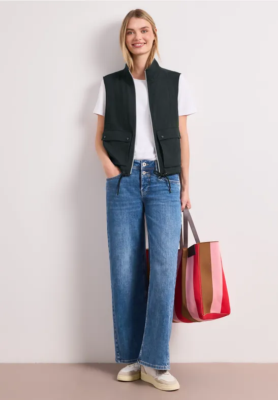 Vrouw met wit T-shirt, zwart vest, rechte blauwe jeans, witte sneakers en rood gestreepte tas