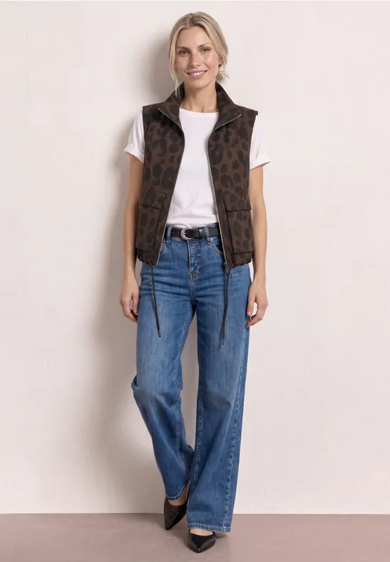 Vrouw in wit T-shirt, bruin mouwloos vest met patroon, blauwe wijd uitlopende jeans en zwarte schoenen.