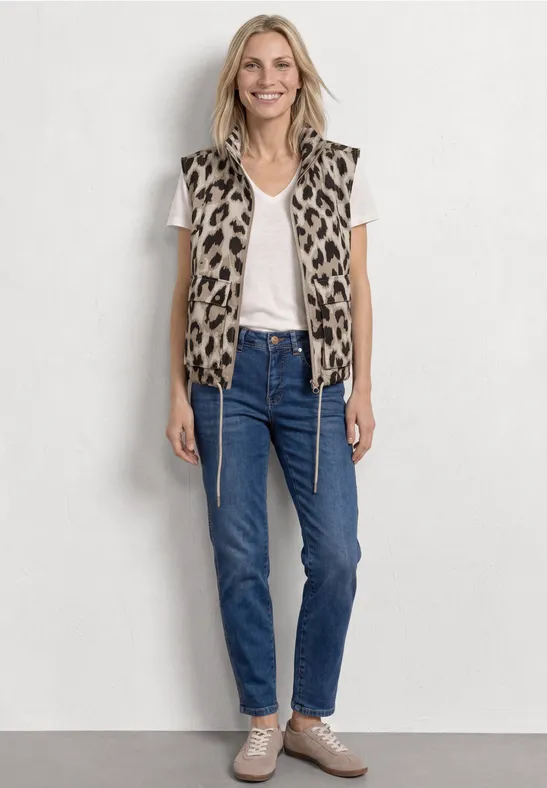 Femme portant un T-shirt blanc, une veste léopard, un jean bleu et des baskets beiges, debout devant un mur clair.