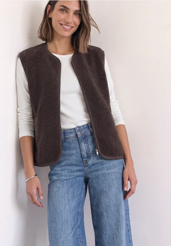 Frau trägt weiße Bluse, braune Fleeceweste und blaue Jeans, steht lächelnd vor hellem Hintergrund.
