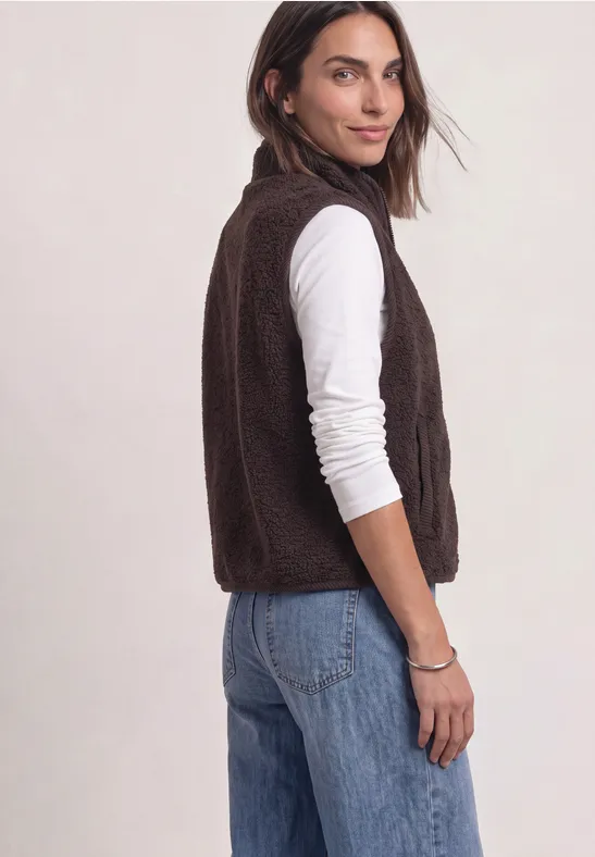 Frau in ¾-Ansicht trägt braune Fleece-Weste, weißes Langarmshirt und blaue Jeans vor hellem Hintergrund.
