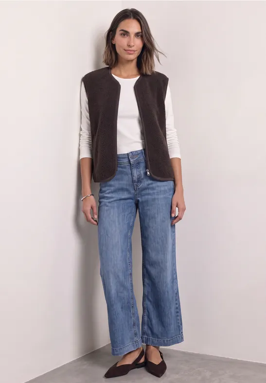 Eine Frau vor heller Wand trägt weiße Bluse, braune Weste, weite Jeans und schwarze Ballerinas.