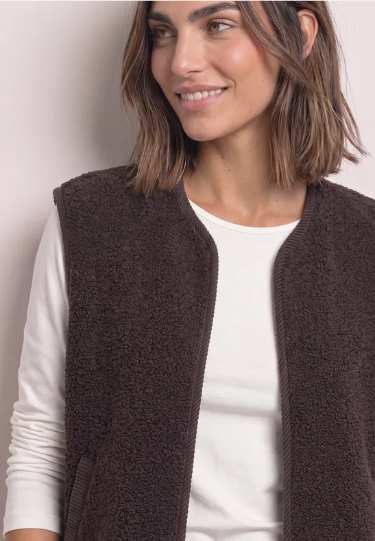 Eine lächelnde Frau trägt einen braunen Teddy-Fleece-Weste über einem weißen Langarmshirt.