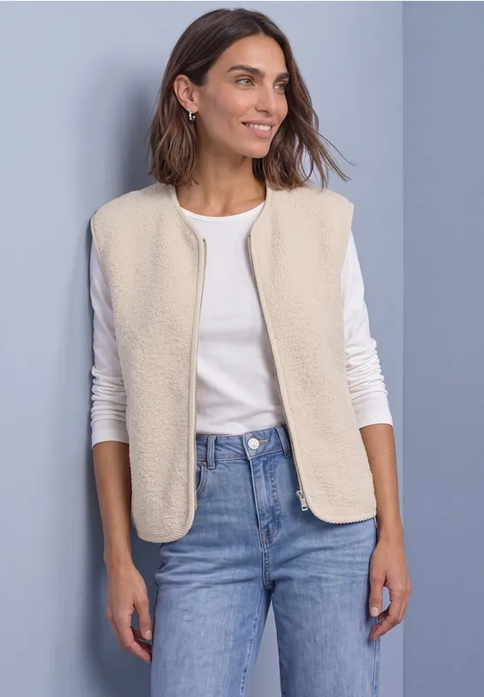 Eine Frau in weißem Langarmshirt, beigefarbener Fleeceweste und Jeans vor hellblauem Hintergrund.