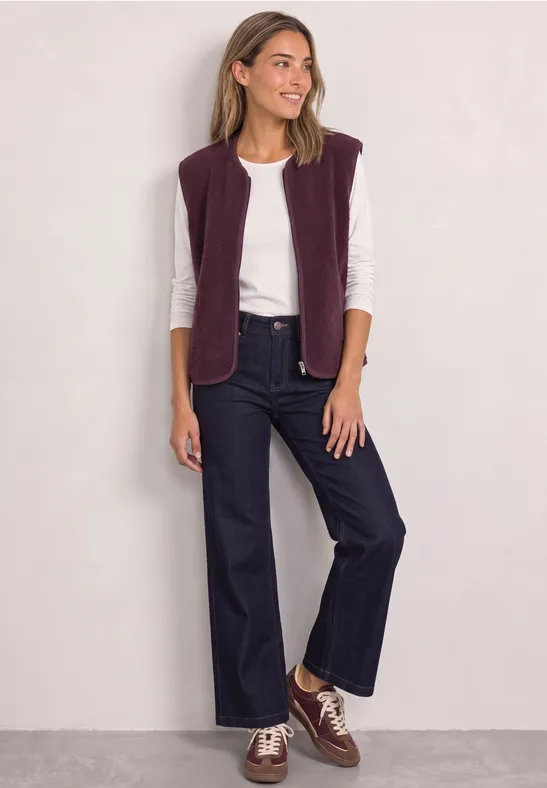 Frau in weißem Shirt, lila Weste, weiten Jeans und braunen Schuhen vor hellem Hintergrund, lächelnd.