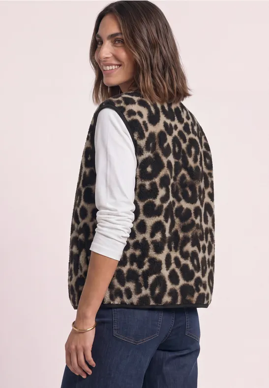 Frau in Animal-Print-Weste und weißem Shirt lächelt seitlich, trägt Jeans.