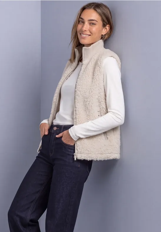 Femme en pull col roulé blanc et gilet polaire beige devant un fond gris.